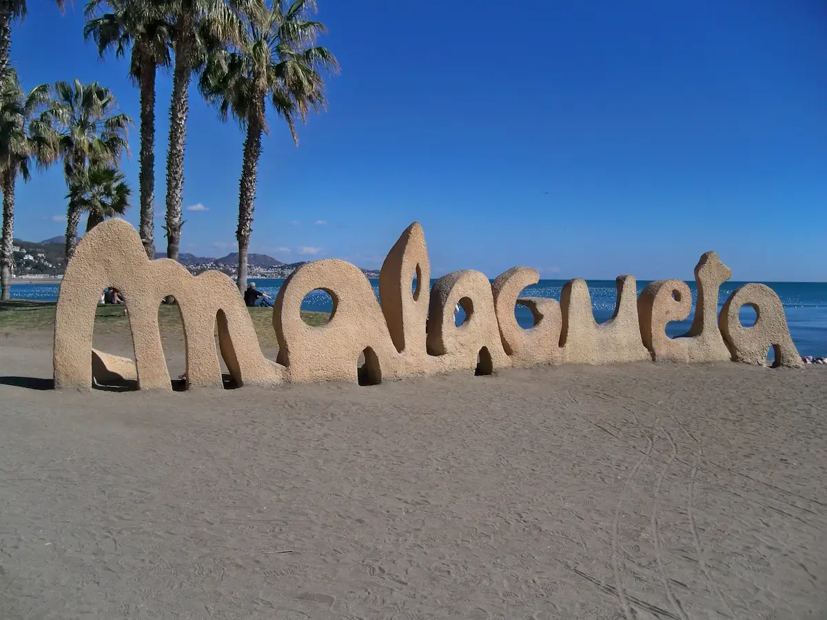 Playa de la Malagueta in Malaga on a sunny day, with palm trees, sunbathers, calm Mediterranean waters, and the city skyline in the background.