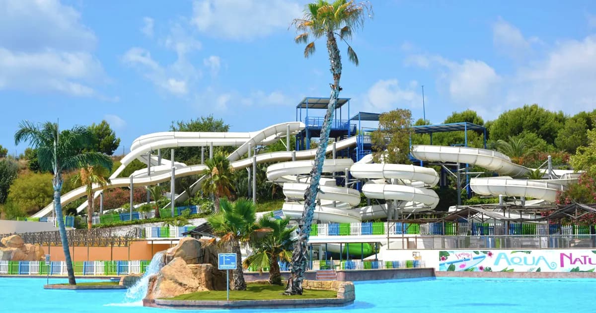 Aerial view of Aqualandia waterpark in Benidorm, Costa Blanca, Spain – family resort with white twisting waterslides, blue pools, palm trees, and colorful Aqua Natura sign under sunny skies.