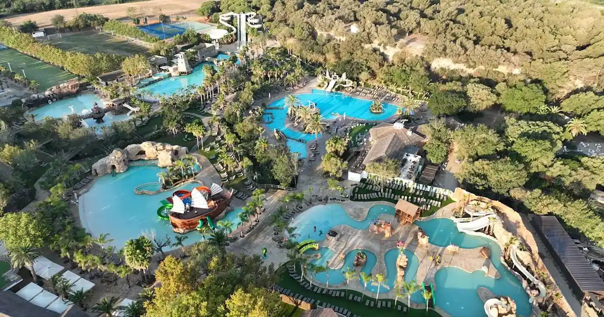 Aerial drone view of large family waterpark resort on Costa Brava, Spain: blue pools, twisty waterslides, pirate ship playgrounds, palm trees, lush green hills.