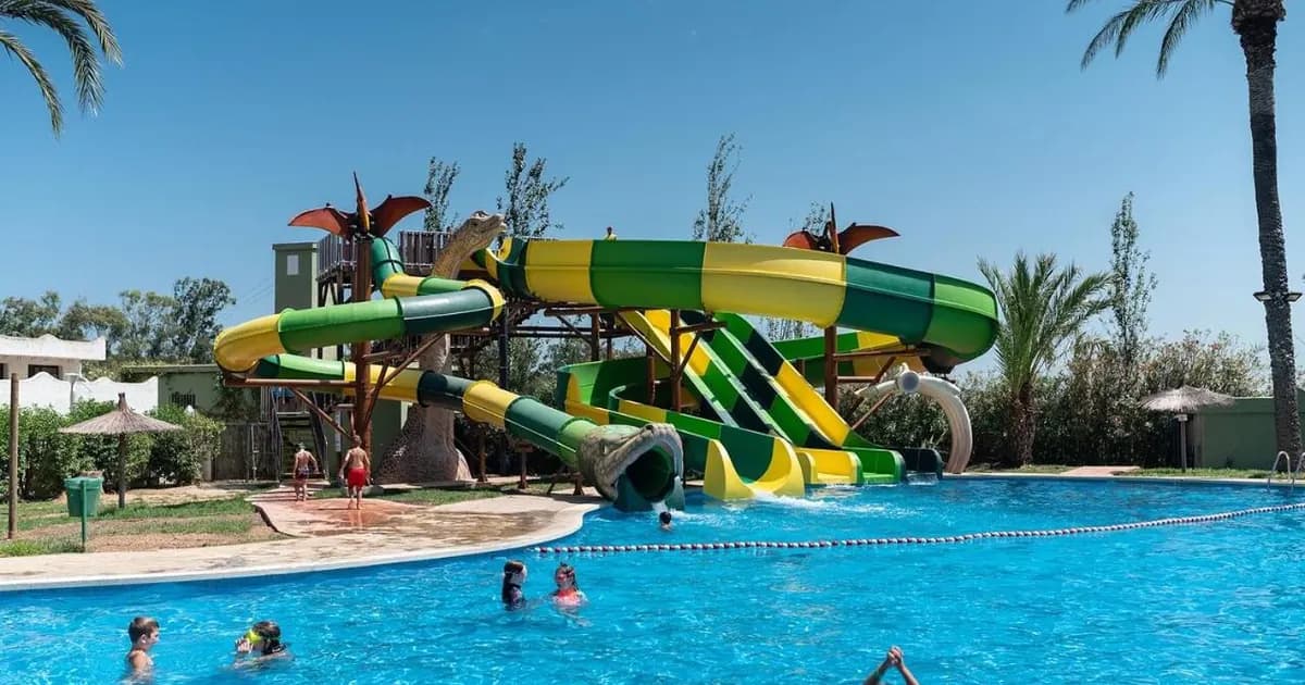 Family waterpark resort on Costa Valencia, Spain: kids playing in blue pool under green-yellow dragon waterslides, palm trees, sunny skies