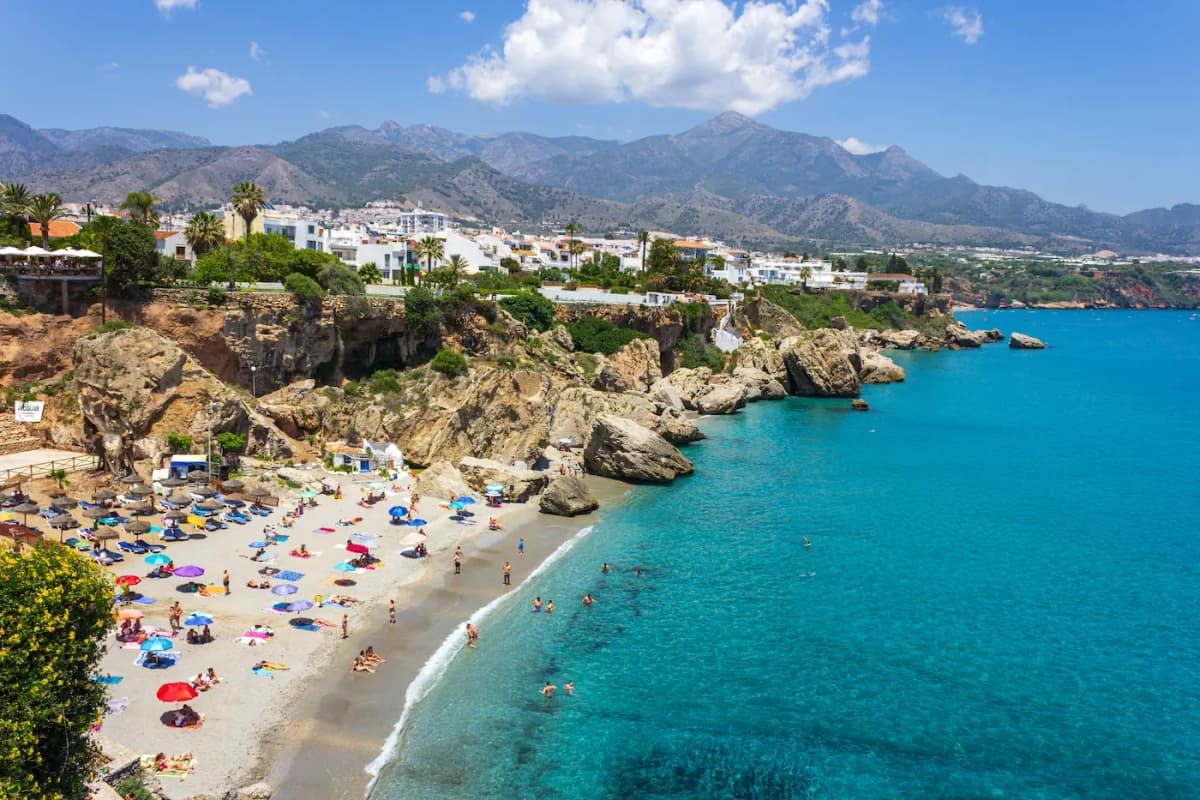 Turquoise sea and sandy beach with umbrellas, rocky cliffs, and mountains on the Nerja