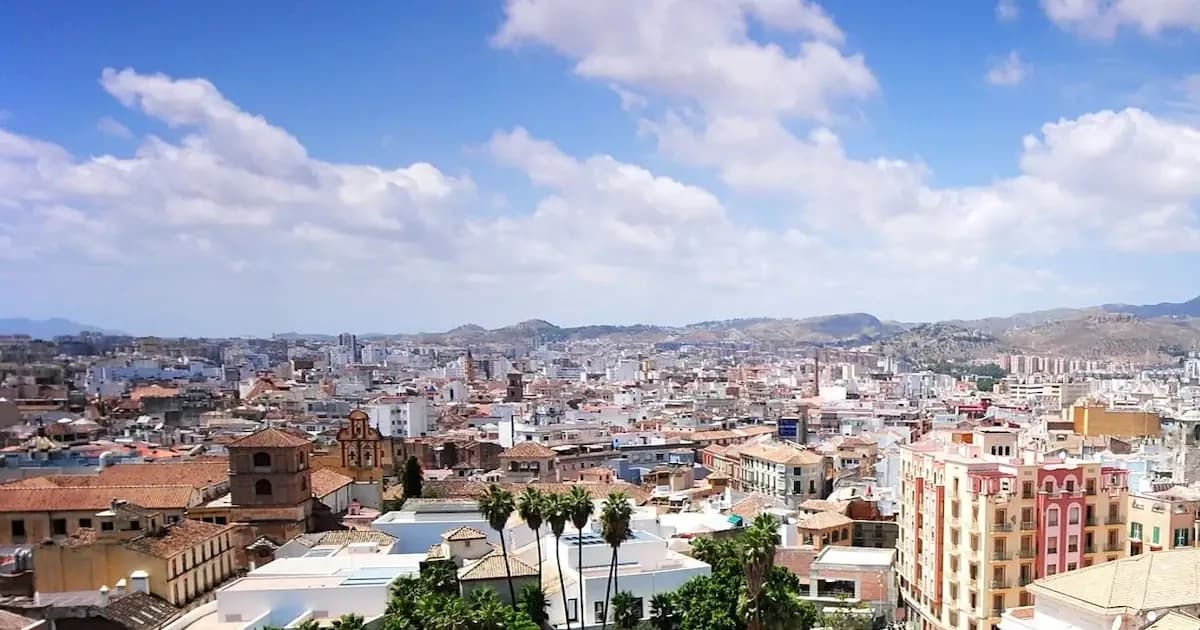 Panoramic view of torremolinos on the Costa del Sol, featuring dense city buildings, the cathedral, palm trees in the foreground, and mountains in the background under a blue sky with scattered clouds.