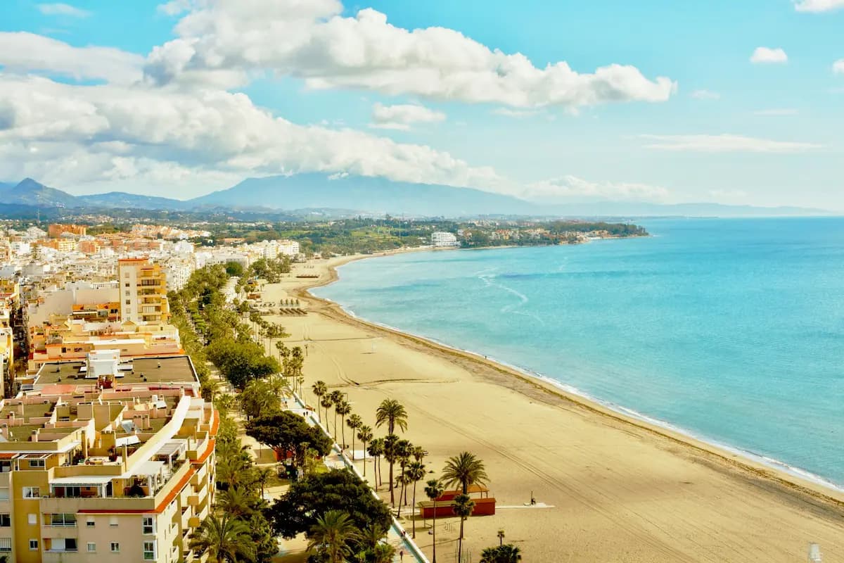 Playa de la Rada in Estepona with blue sky fine grey sand, calm blue Mediterranean sea, palm-lined promenade and beach bars on a sunny day