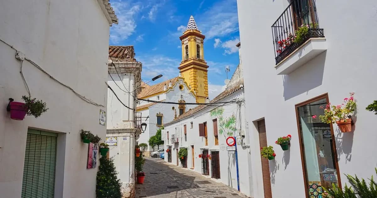 Charming narrow street in Estepona Old Town with whitewashed houses adorned with colorful flower pots, leading to a prominent yellow church tower under blue skies.