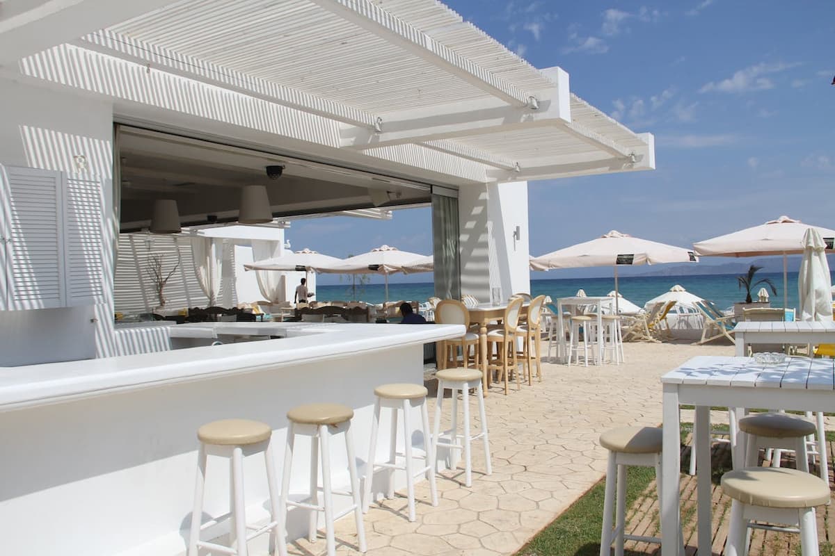 Chic white beachfront restaurant terrace with bar, pergolas, and sea view on a sunny day.