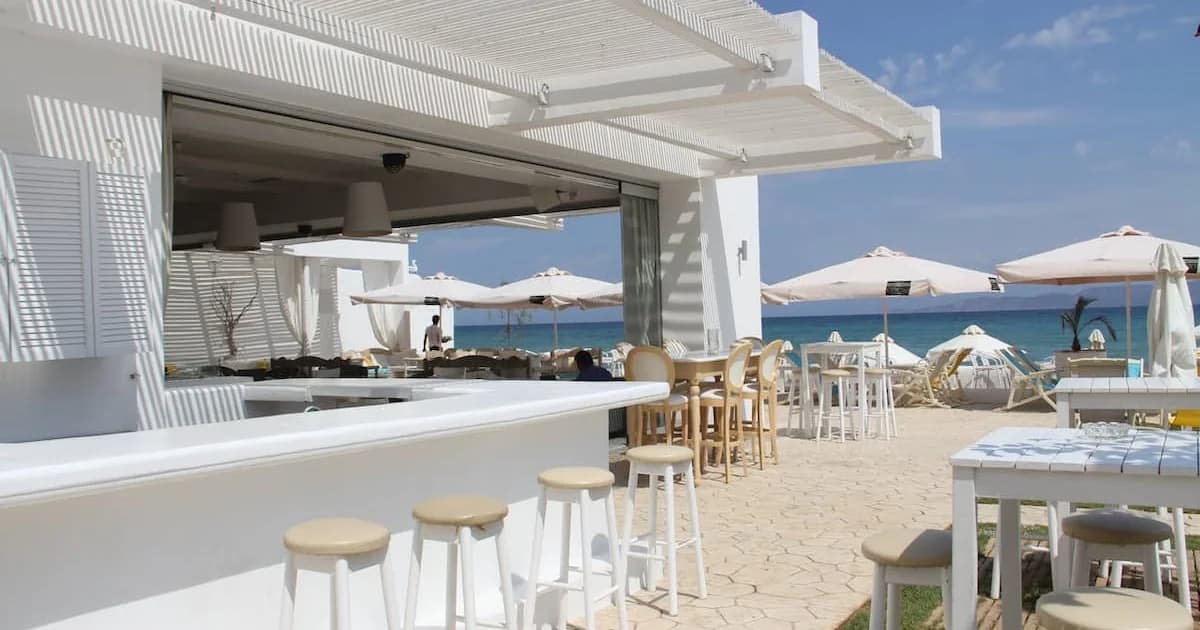 Chic white beachfront restaurant terrace with bar, pergolas, and sea view on a sunny day.