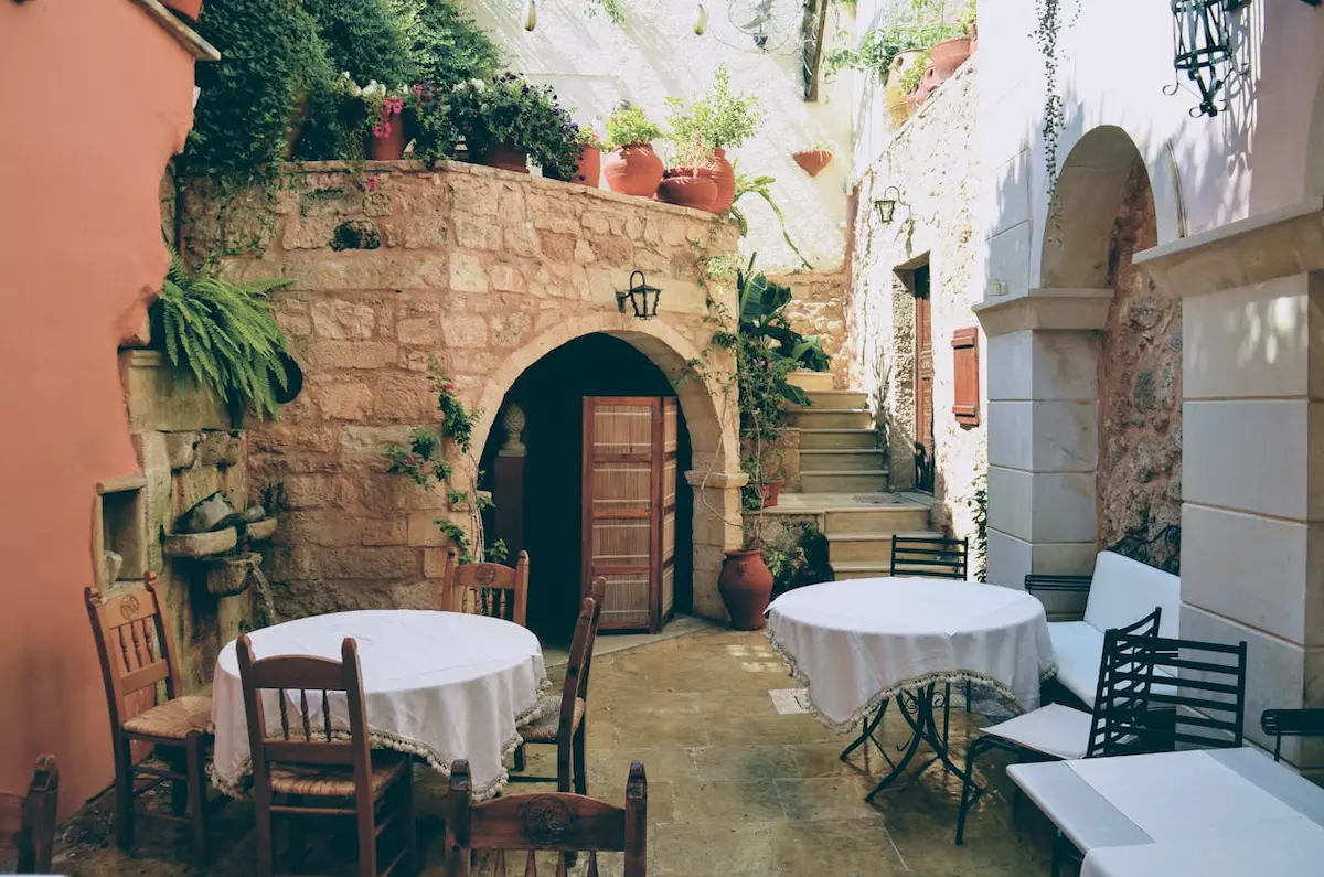 A charming outdoor terrace of a restaurant in Spain, featuring a rustic stone courtyard with arched doorways and walls covered in greenery and potted plants. Wooden tables draped in white tablecloths are set with chairs, ready for al fresco dining under warm lighting.