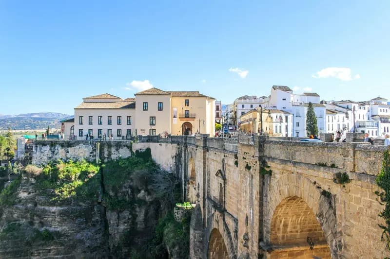 View of Ronda, Spain: the stone arch bridge Puente Nuevo spanning the deep El Tajo gorge, with white and pastel buildings lining the cliff under a bright blue sky.
