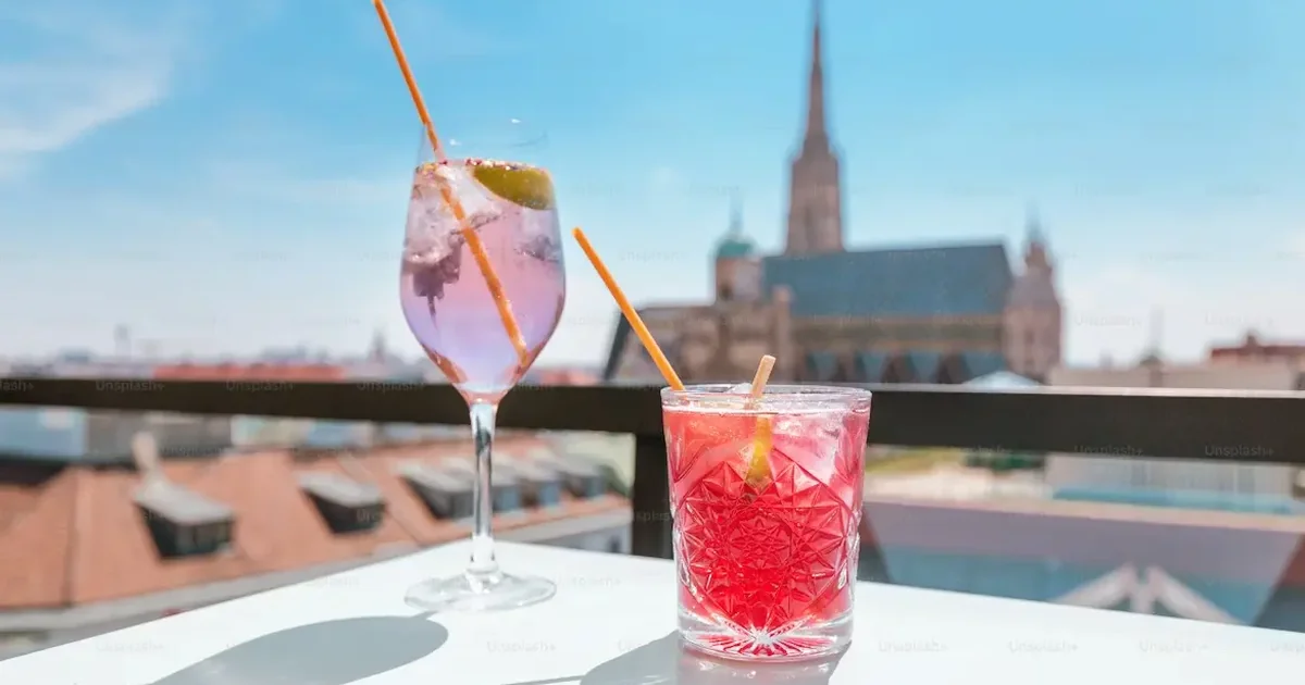 Two vibrant cocktails—a purple gin with lime and a red drink—on a sunny rooftop bar, with a Gothic cathedral spire in the background under blue skies.