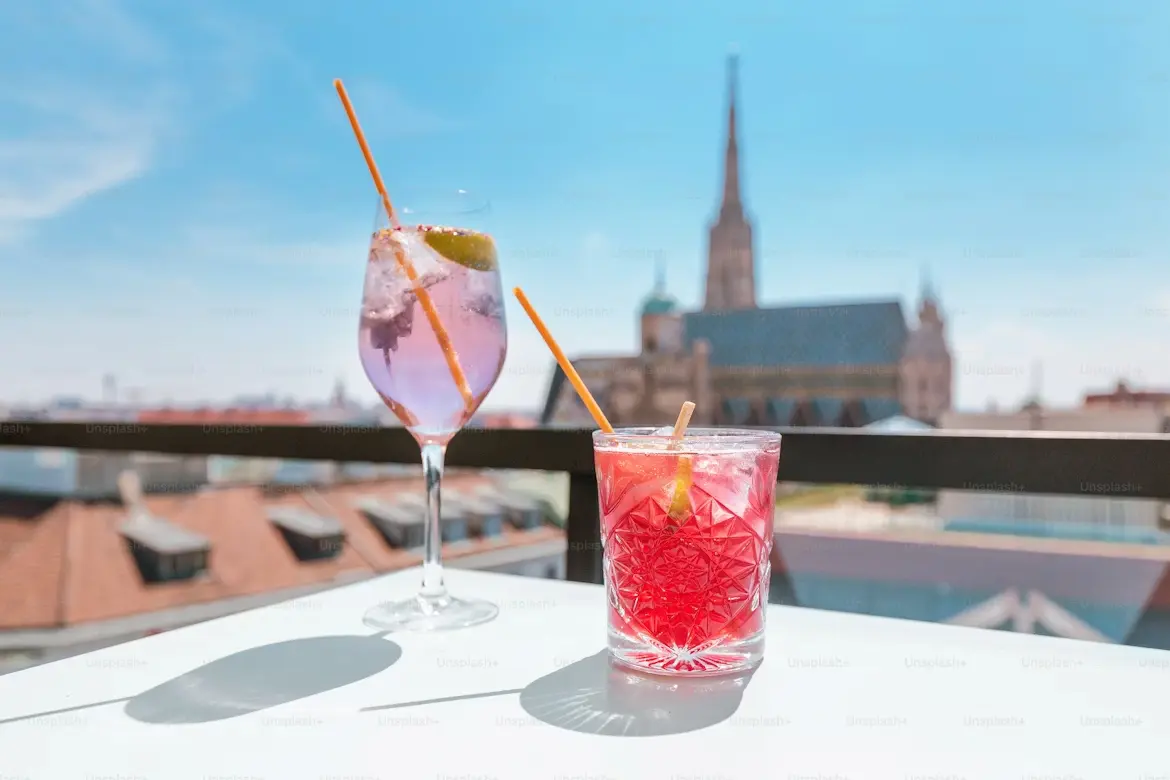 Two vibrant cocktails—a purple gin with lime and a red drink—on a sunny rooftop bar, with a Gothic cathedral spire in the background under blue skies.