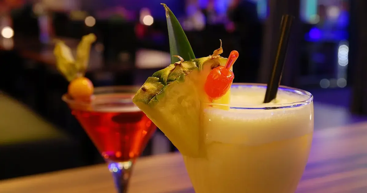 Two tropical cocktails on a wooden bar: yellow piña colada with pineapple slice and cherry, red martini with fruit garnish, purple neon background.