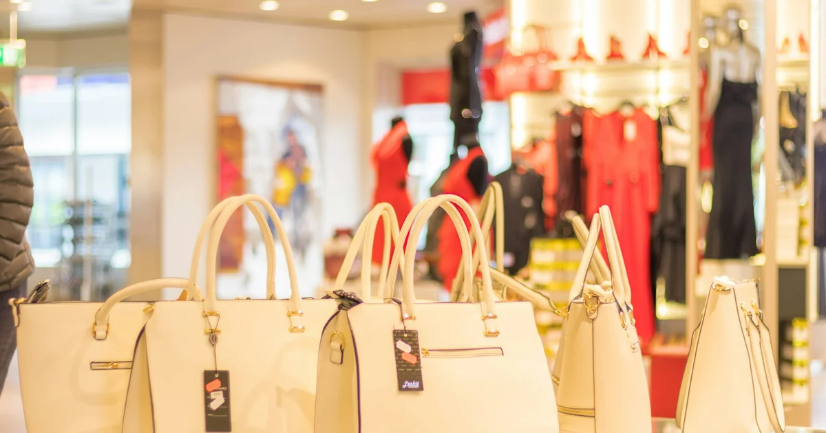 Display of luxury beige handbags in a Malaga shopping centre store, surrounded by women's fashion clothing and accessories.