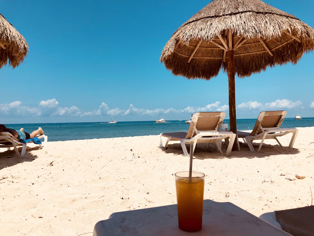 Tropical beach scene with a thatched umbrella, lounge chairs, and a cold drink.