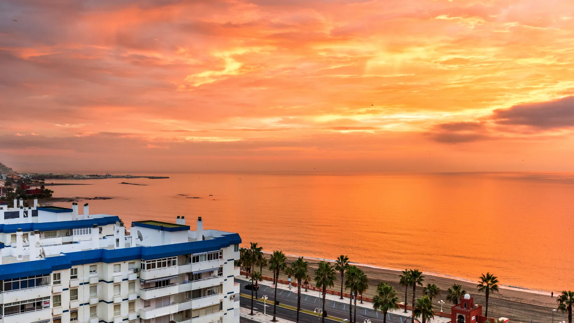 Sunset over the sea in benalmadena with palm-lined promenade and apartment buildings.