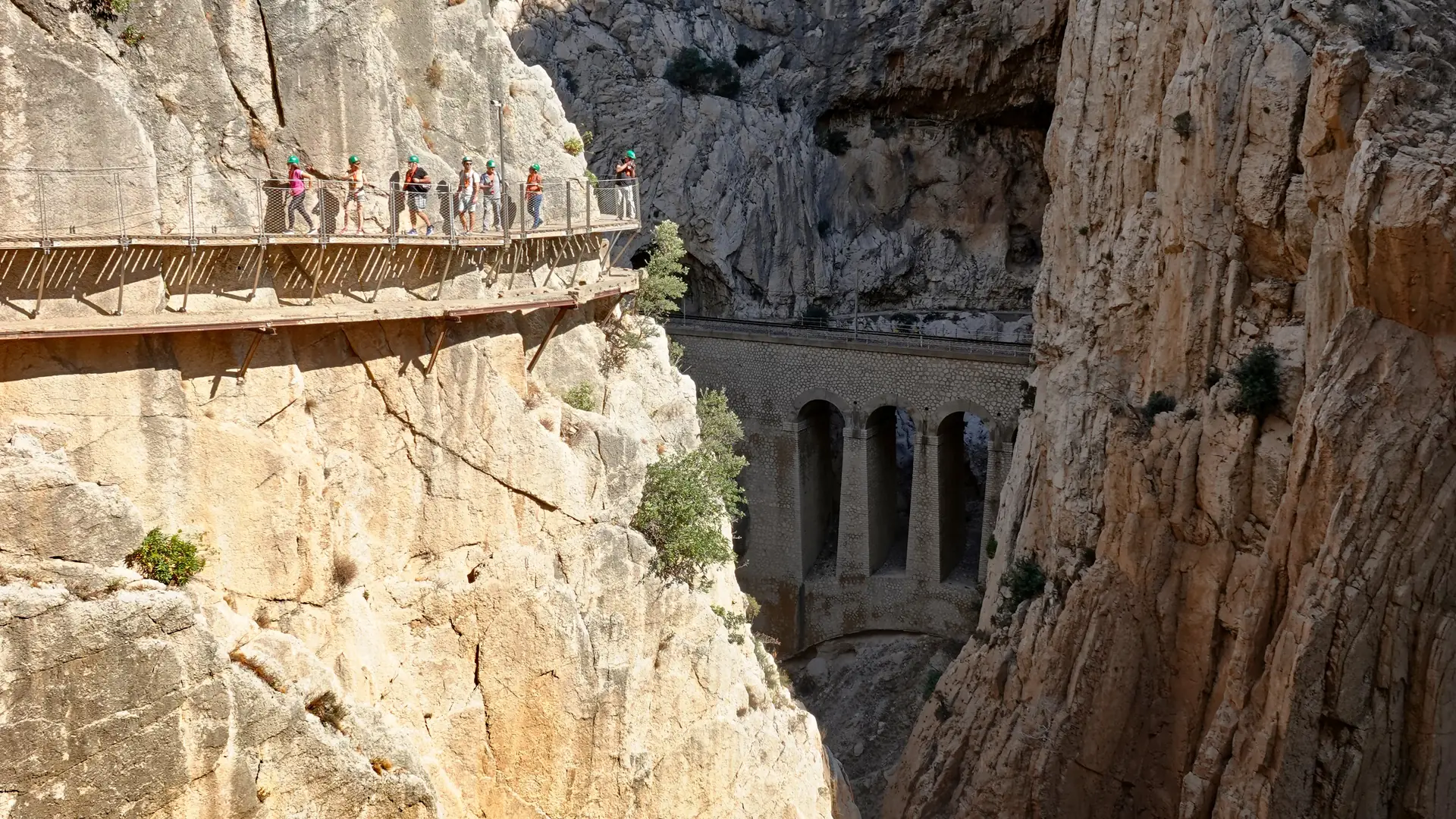 Walkers on the Caminito del Rey boardwalk above the gorge, with the old railway viaduct behind
