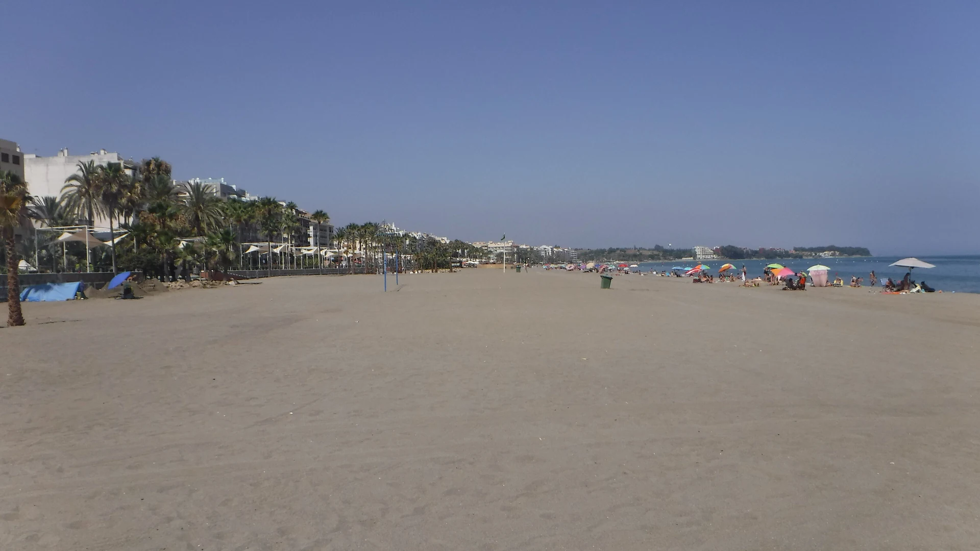 Playa de la Rada in Estepona with fine grey sand, colourful beach umbrellas, a palm-lined promenade and the town behind