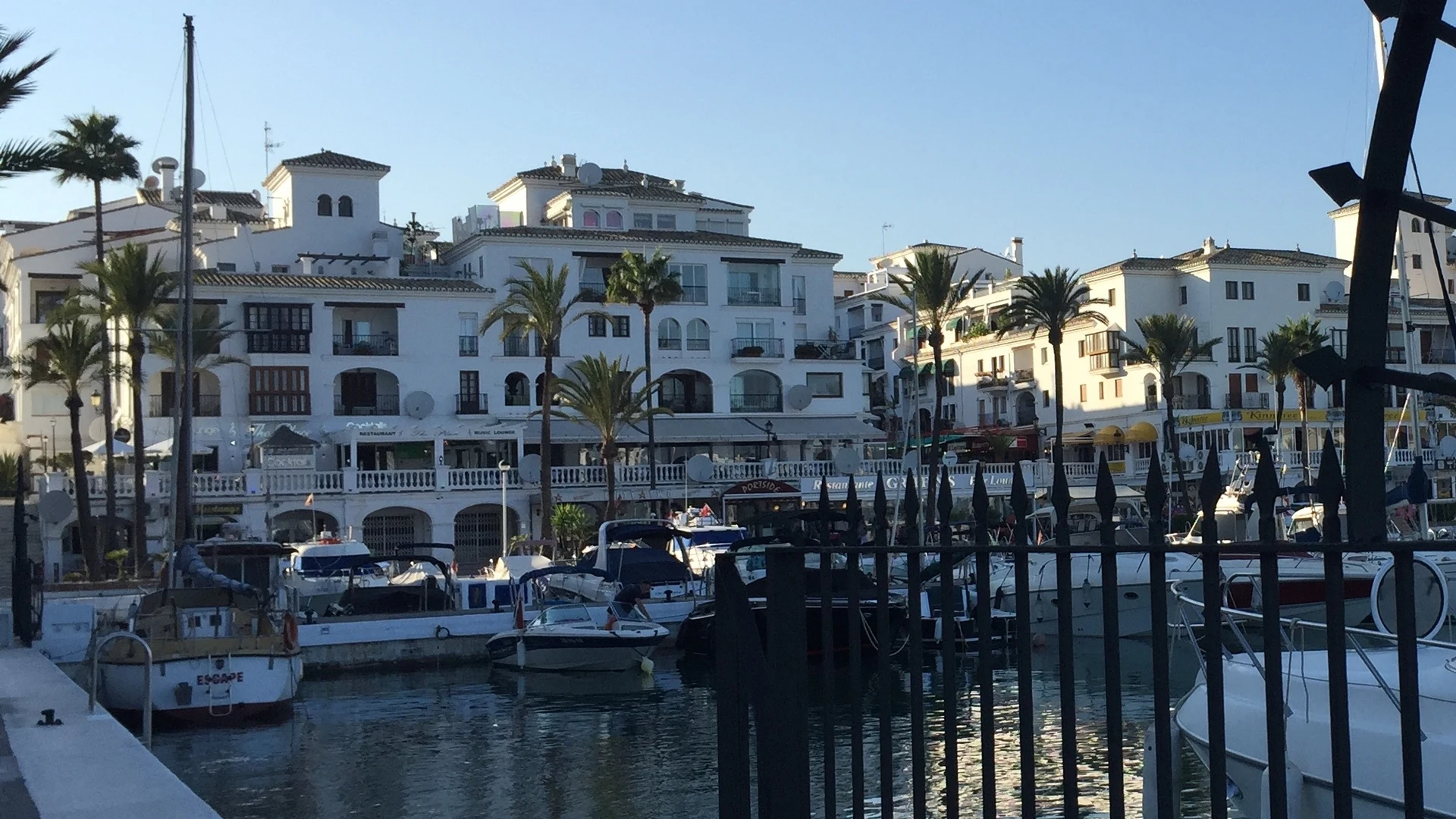 Motor yachts and sailing boats moored in a Costa del Sol marina, with white Andalusian-style apartments and palm trees along the promenade